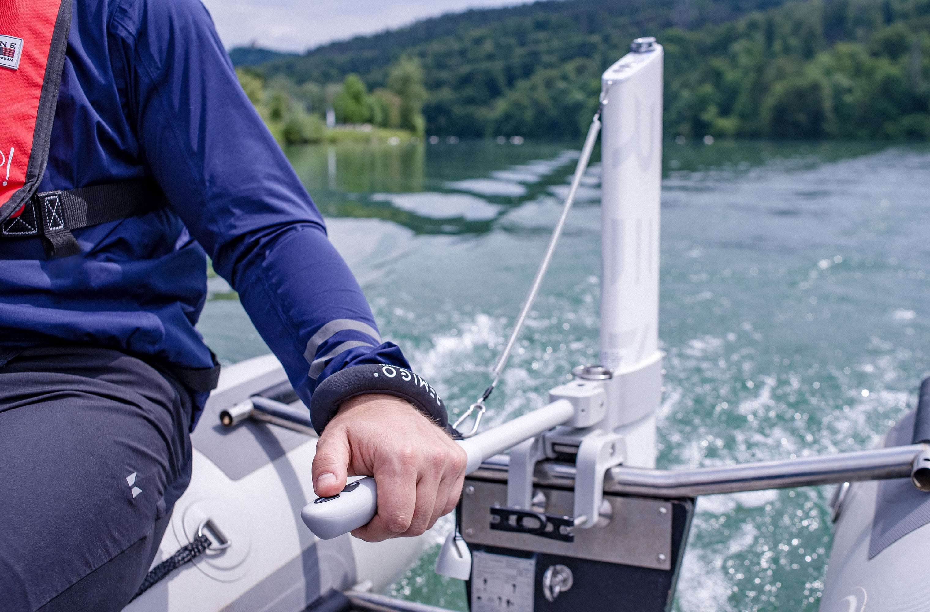 Person on a boat holding onto rudder extension with remote control with scenic lake and trees in the background